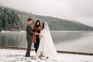 Winter lakeside elopement ceremony with couple exchanging vows before an officiant on snowy shore.