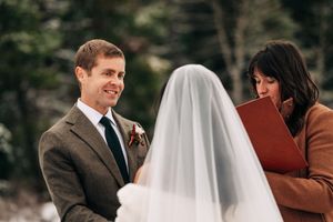 Groom smiling during winter wedding ceremony as officiant reads from red book beside snowy trees.