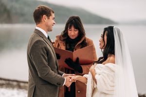 Bride and groom hold hands during intimate winter lakeside wedding ceremony with officiant.