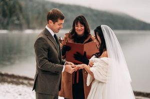 Groom places wedding ring on bride's finger during outdoor winter ceremony beside a misty lake.