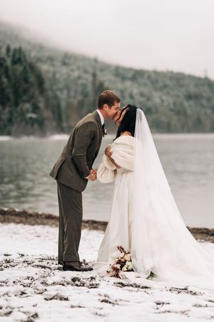 Bride and groom share first kiss on snowy lakeshore with misty forested mountains behind.