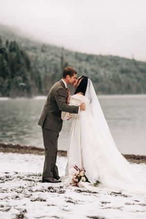 Bride and groom share romantic kiss by snowy lakeside with misty forested mountains backdrop.