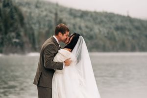 Bride and groom share a romantic kiss by a serene snowy lake during their winter wedding celebration.