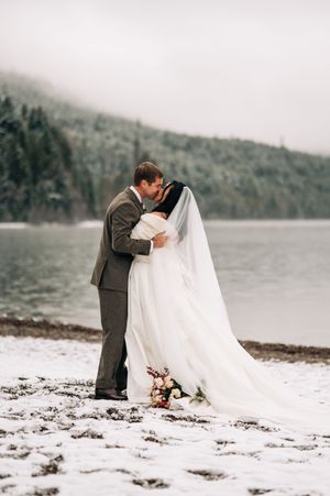 Newlyweds share first kiss on a snowy lakeshore with misty forested mountains in the background.