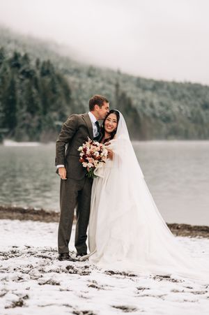 Groom kisses bride's cheek as she smiles holding bouquet by snowy lake at winter wedding.