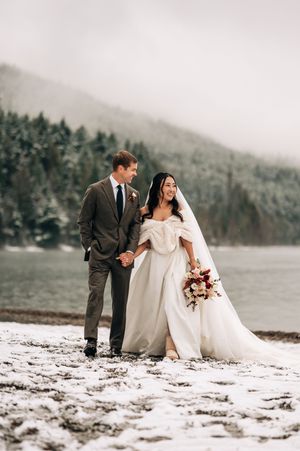 Bride and groom holding hands on snowy lakeside with snow-dusted evergreen forest backdrop.