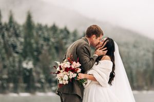Newlyweds share a romantic kiss holding a red and pink bouquet in a snowy winter setting.