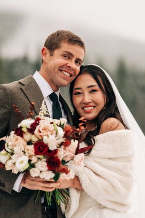 Happy couple poses together holding red and pink floral bouquet at snowy winter wedding.
