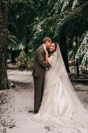 Bride and groom embrace in a snowy winter forest, bride wearing a flowing white gown and long veil.