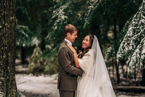 Couple embracing in snowy winter forest, bride in white gown and veil, groom in brown suit, surrounded by snow-dusted trees.