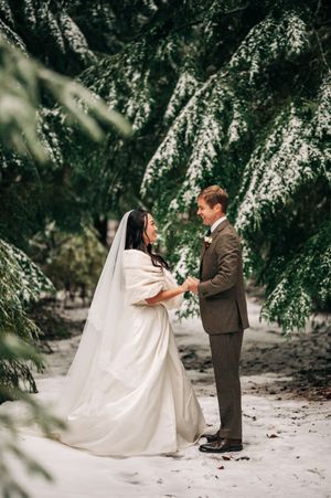 Bride and groom hold hands facing each other among snow-dusted evergreen trees in a winter wedding ceremony.
