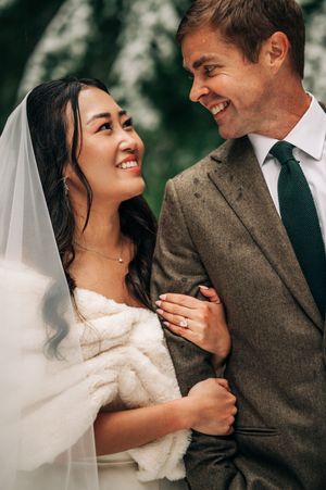 Bride and groom smiling at each other outdoors in winter, she in white gown and veil, he in grey tweed suit.