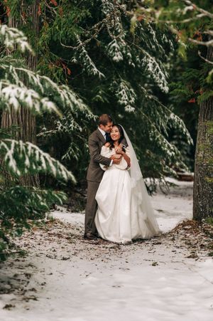 Groom embraces bride from behind as they smile together on a snowy forest path lined with pine trees.