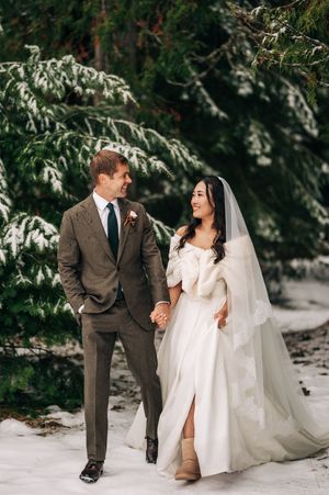 Smiling bride in fur wrap and groom in brown suit walk hand in hand through a snowy evergreen forest.