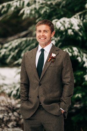 Smiling groom in brown tweed suit with boutonniere poses outdoors in snowy winter wedding setting.