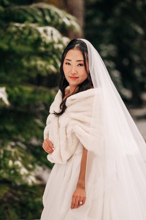 Elegant bride in white fur stole and cathedral veil poses before snow-covered evergreen trees outdoors.