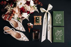 Elegant wedding flat lay with lace heels, burgundy bouquet, perfume, and green vow books on black.