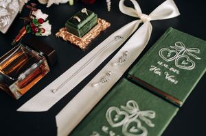 Elegant wedding flat lay with green vow books, white ribbon, ring box, and perfume.