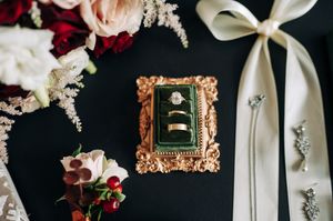 Wedding rings displayed in ornate gold velvet box surrounded by red and blush floral arrangements.