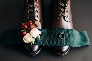 Brown leather lace-up boots with a teal tie, gold wedding ring, and floral boutonniere on dark background.