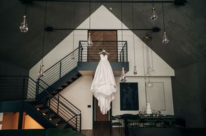 Elegant white wedding gown hanging in modern industrial venue with exposed stairs and pendant lights.