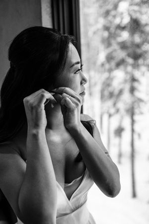 Black and white photo of bride putting on earrings by window on wedding day.