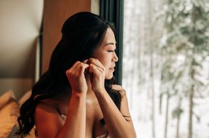 Bride puts on earrings by a frosted window with snowy trees visible in the background.