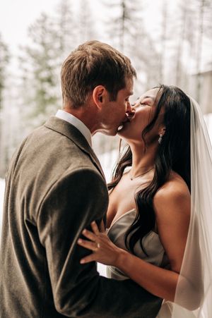 Bride and groom share a romantic kiss outdoors surrounded by tall pine trees on wedding day.