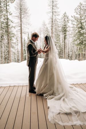 Bride and groom exchange rings on snowy winter wedding deck surrounded by snow-covered pine trees.