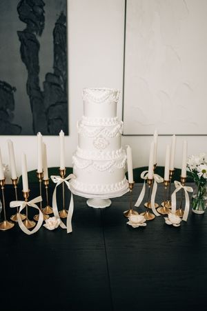 Elegant three-tier white wedding cake with pearl and floral details, surrounded by gold candlesticks with ribbon bows.