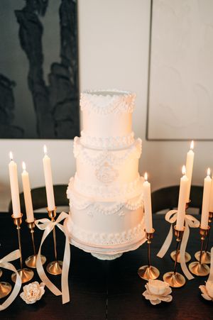 Elegant four-tier white wedding cake with lace details surrounded by gold candlesticks and ribbon bows.