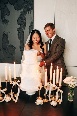 Bride and groom cutting a white tiered wedding cake surrounded by lit candles on a dark table.