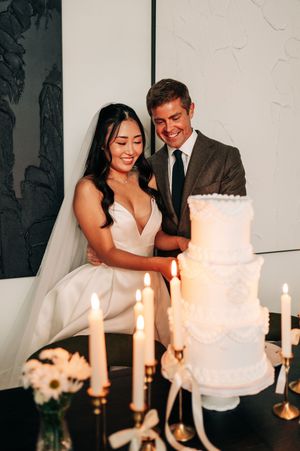Smiling bride and groom cut their white tiered wedding cake surrounded by glowing candlelight.
