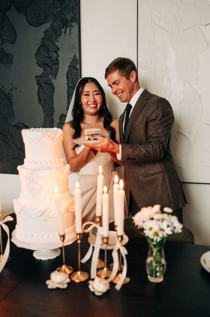 Smiling newlyweds holding a slice of wedding cake beside glowing candles and white floral arrangements.
