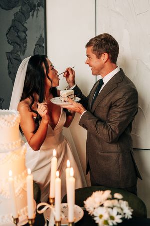 Groom feeds bride a slice of wedding cake by candlelight, bride in white gown with veil, groom in gray suit.