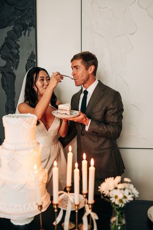 Bride feeds groom wedding cake slice at candlelit reception table with tiered white cake.