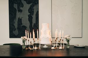 Elegant white tiered wedding cake surrounded by gold candlesticks and florals on dark table.