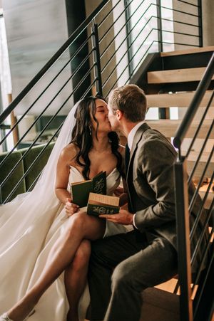 Bride and groom share a kiss on modern staircase holding green wedding vow books.