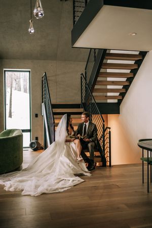 Bride and groom sit together on modern staircase inside contemporary venue with snowy view outside.