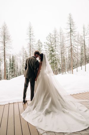 Bride and groom sharing a kiss on a snowy deck surrounded by snow-covered pine trees in winter.