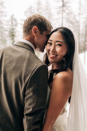 Smiling Asian bride in white dress and veil leans into groom in gray suit in winter setting.