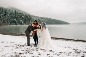 Officiant performs winter wedding ceremony for couple beside snow-covered lake with foggy mountains.