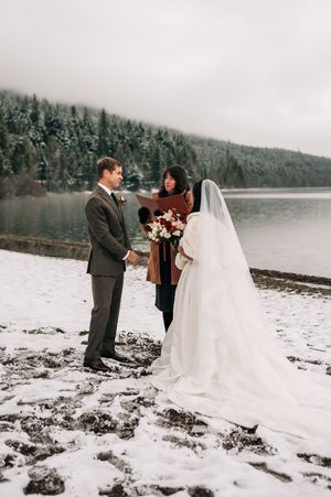 Outdoor winter wedding ceremony by a misty lake with snow-covered ground and evergreen forest backdrop.