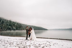 Couple exchanging vows on snowy lakeside shore with evergreen forest and misty mountains in winter wedding ceremony.
