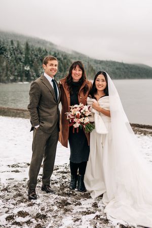 Bride in white gown with bouquet, groom in gray suit, and officiant pose by snowy lakeside.