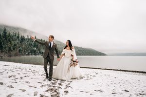 Joyful couple walks hand-in-hand along snowy lakeshore with misty forested mountains behind them.