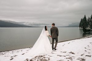 Bride and groom hold hands gazing at scenic snow-covered lake and mountain landscape together.