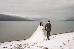 Bride and groom holding hands walking along snowy lakeside with misty mountain backdrop.