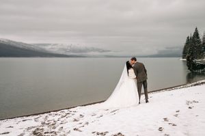 Bride and groom share a kiss on a snowy lakeside shore with misty mountains in the background.