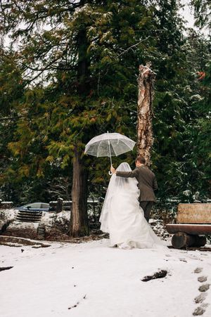 Bride and groom walk under a clear umbrella through a snow-covered forest in winter wedding attire.